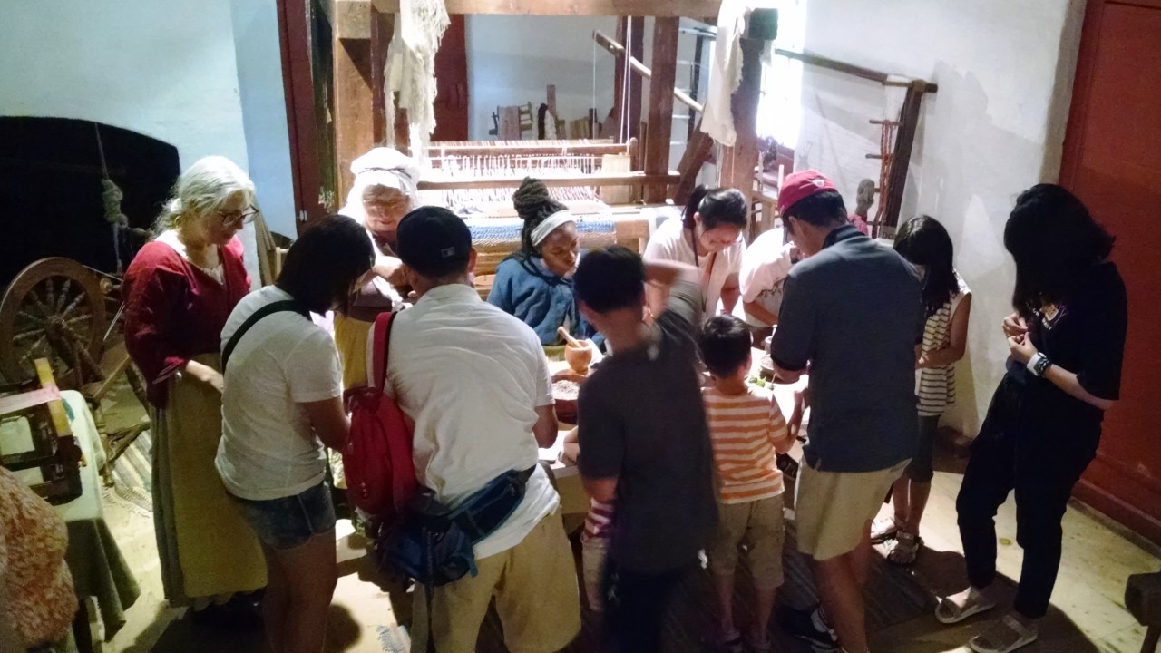 A group of children and adults lean over a table with two costumed interpreters showing them how to make potourri