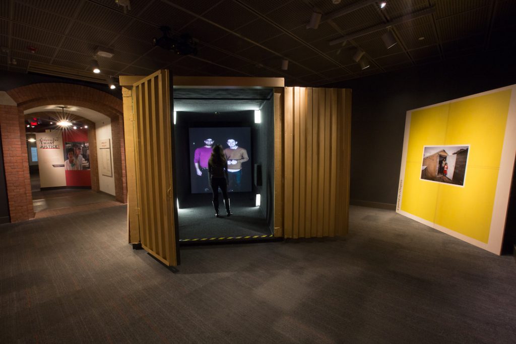 A shipping container painted yellow sits in a gallery with a video screen at the end where two individuals are shown on the screen while another interacts with them in real life.