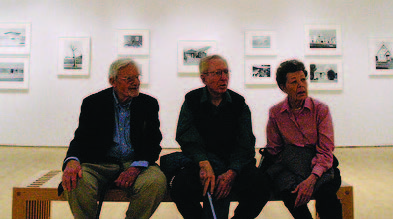 Three older adults (two men, one woman) sit on a bench in a gallery