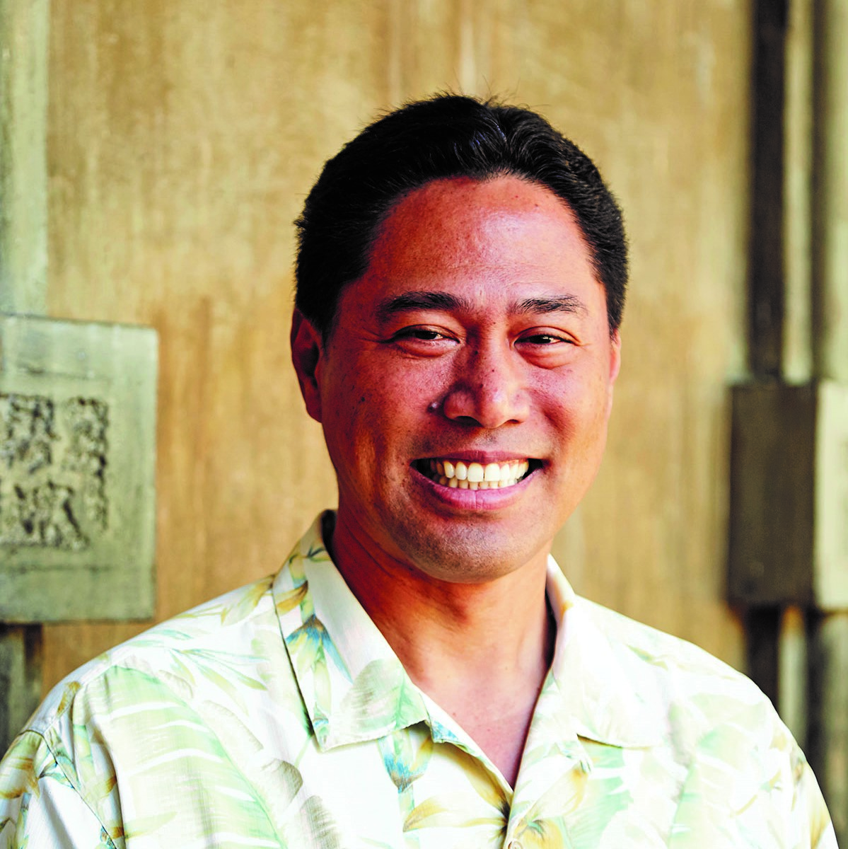 A native man stands in front of a concrete wall wearing a Hawaiian style shirt. 