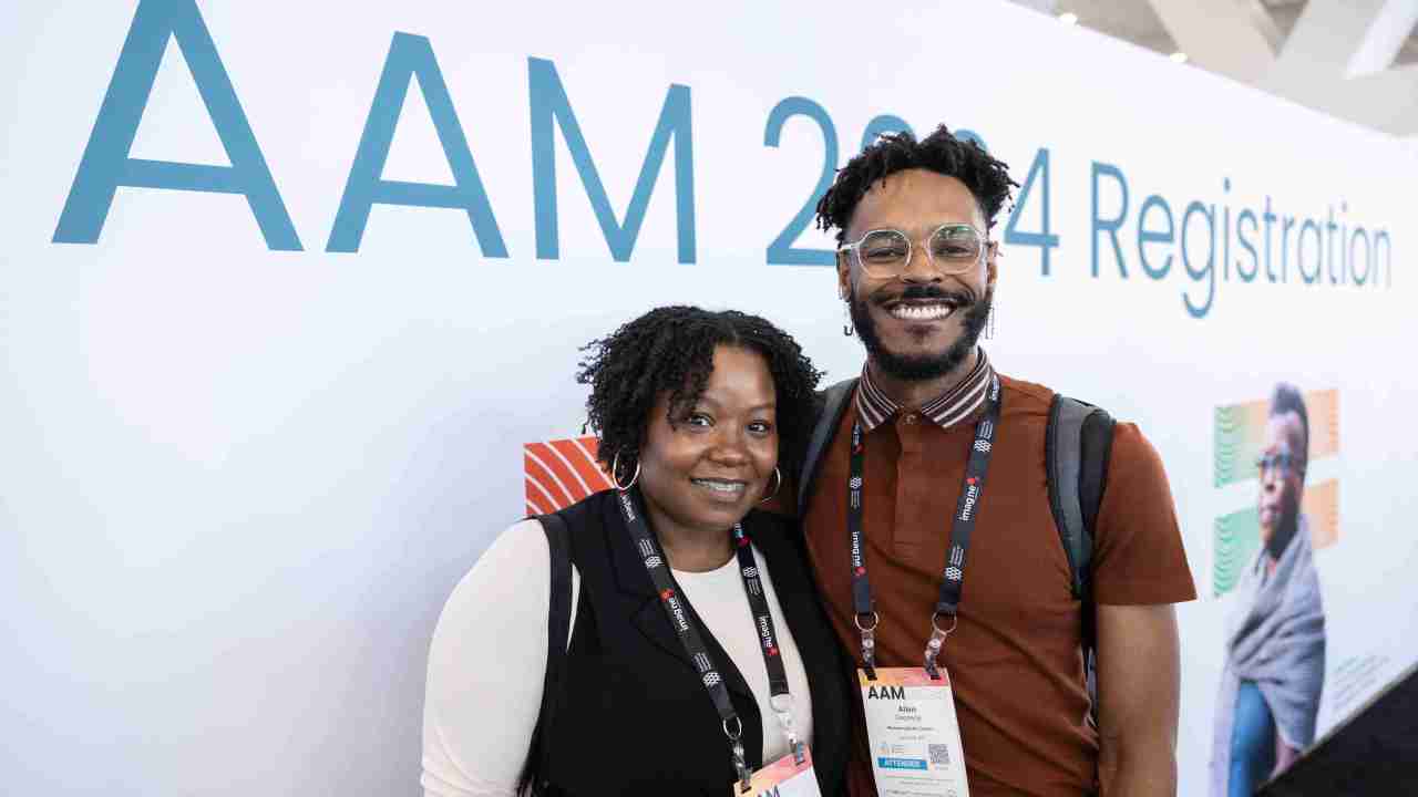 Two people posing for a photo in front of the AAM 2024 registration lobby.