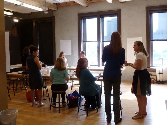 people are seated around a conference table with sunlit windows in the background. Two people stand talking to each other in the foreground