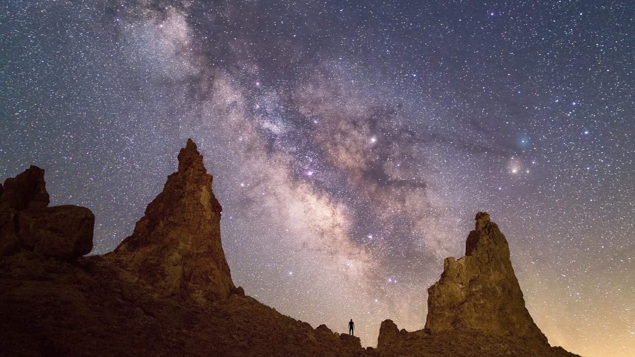 Image of the night sky with several sharp peaks in the foreground and a small person standing.