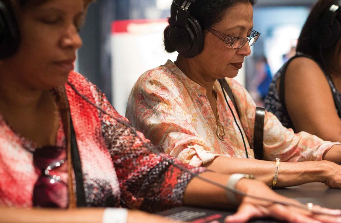 Two women sit at a grey countertop one is clearly wearing headphones both have their hands on the counter looking down.