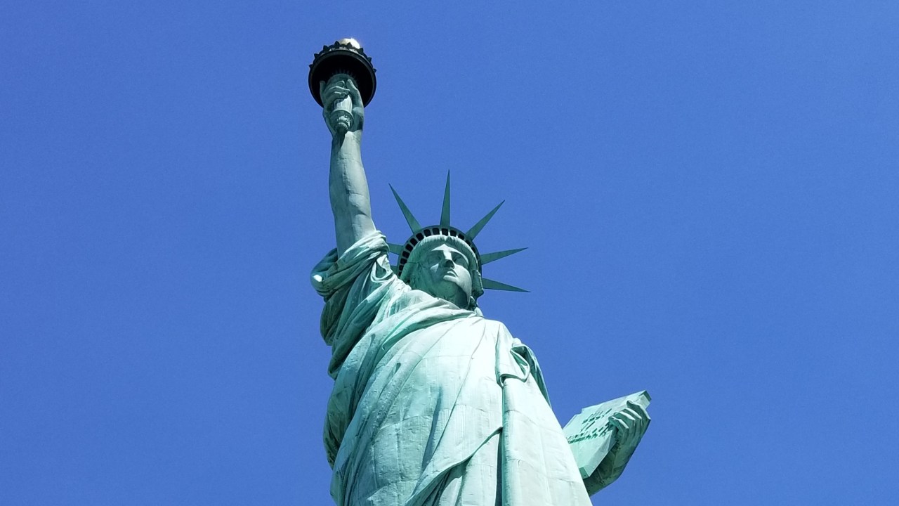 Image of the top of the Statue of Liberty taken from below looking up with a clear blue sky behind her patina-ed green.