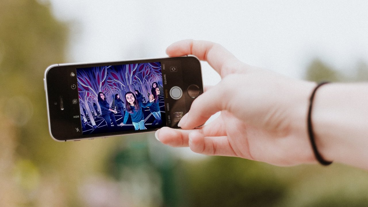 A person's hand holds a smartphone displaying a photograph of one child and one adult frolicking in a mirrored art installation that appears to stretch into infinity, an example of the sort of installations that attract outsize attendance and social media pickup.