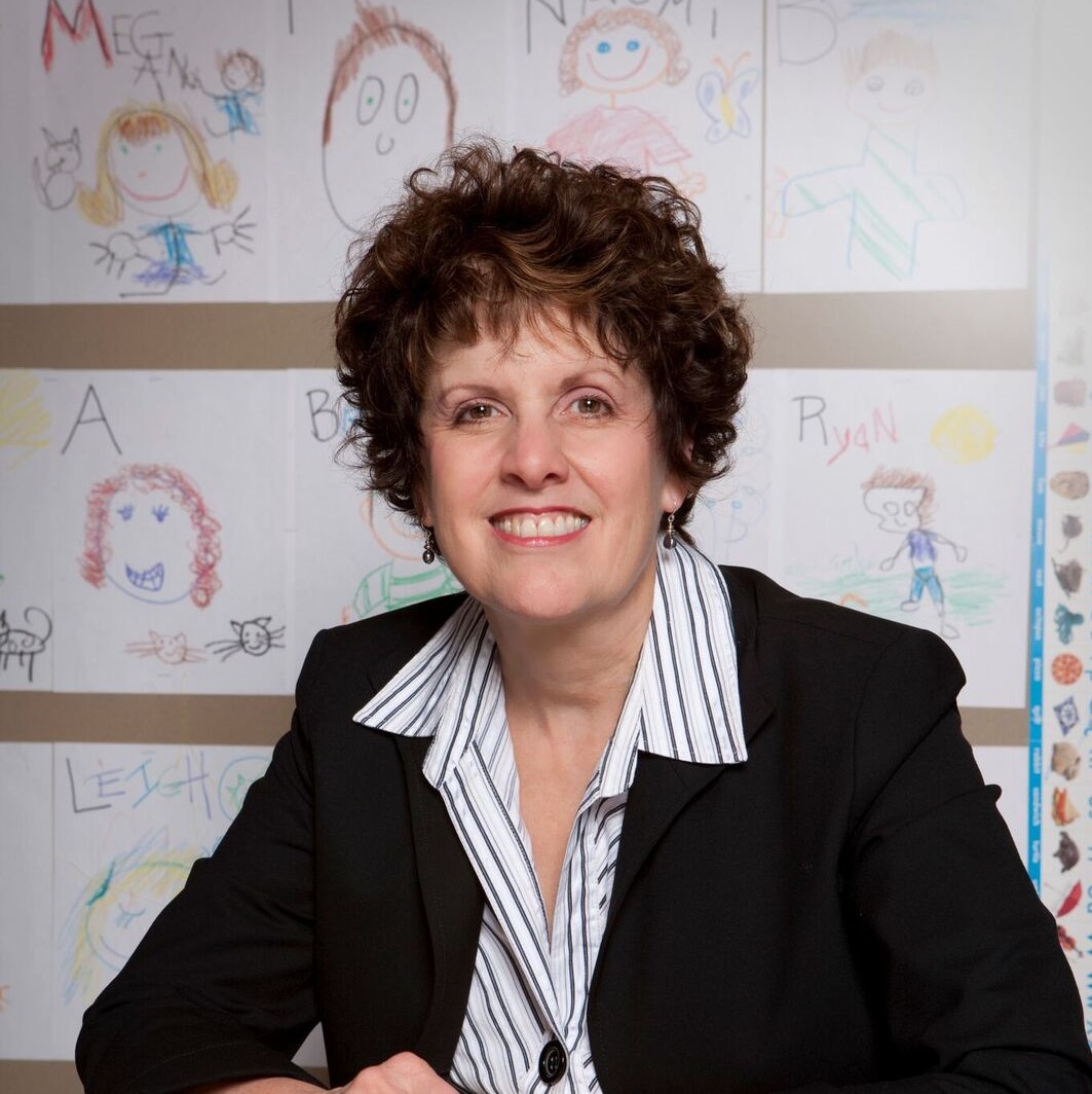 Image of Carole Charnow, a white woman sitting in front of a wall with children's drawings wearing a dark suit jacket and striped shirt. 