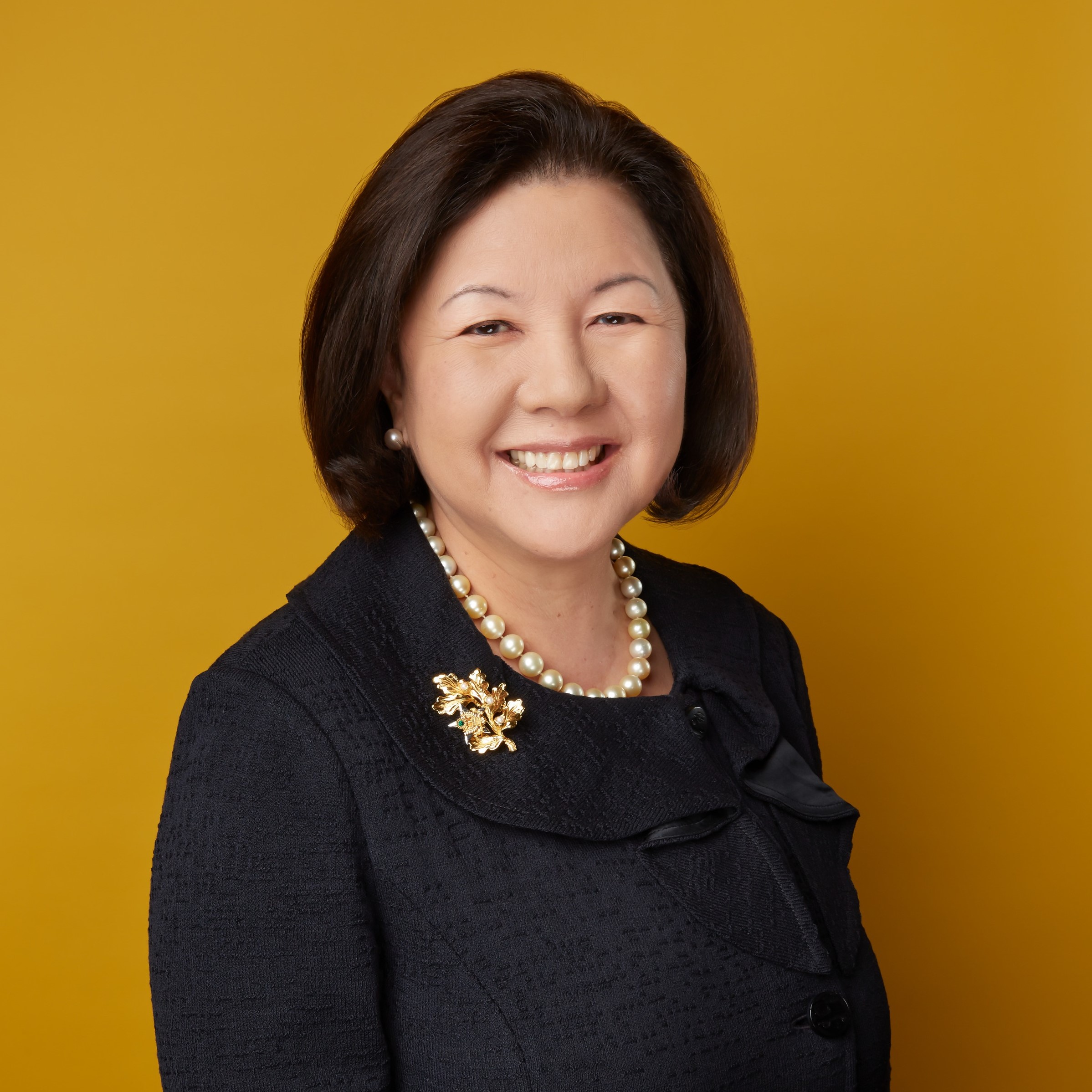 Image of Irene Hirano, an Asian woman standing in front of a mustard colored wall wearing a dark grey dress and pearls. 