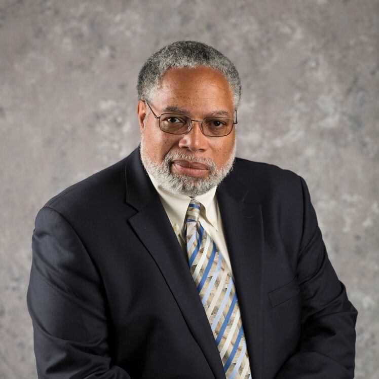 Image of Lonnie Bunch, a black man with silver and black hair, sits in front of a grey backdrop wearing a dark grey suit with a striped tie and metal framed glasses.