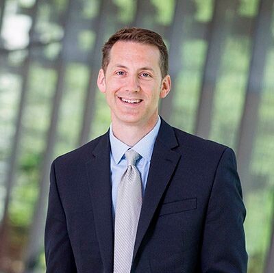 Image of Rod Bigelow, a white man standing in front of a tree backdrop wearing a dark suit and light colored tie. 