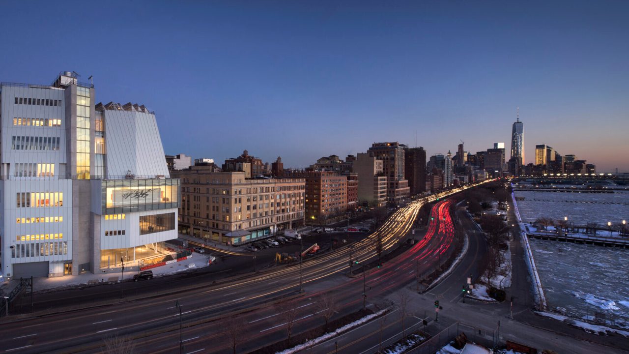 An exterior of the Whitney Museum building at sunset, with a view of the Hudson River nearby