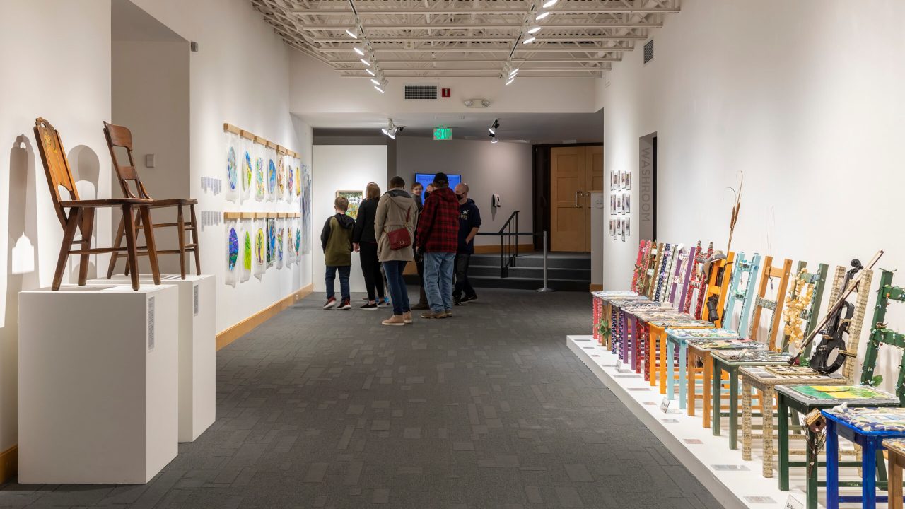 A gallery of decorative chairs and wall hangings with a huddle of visitors looking at them