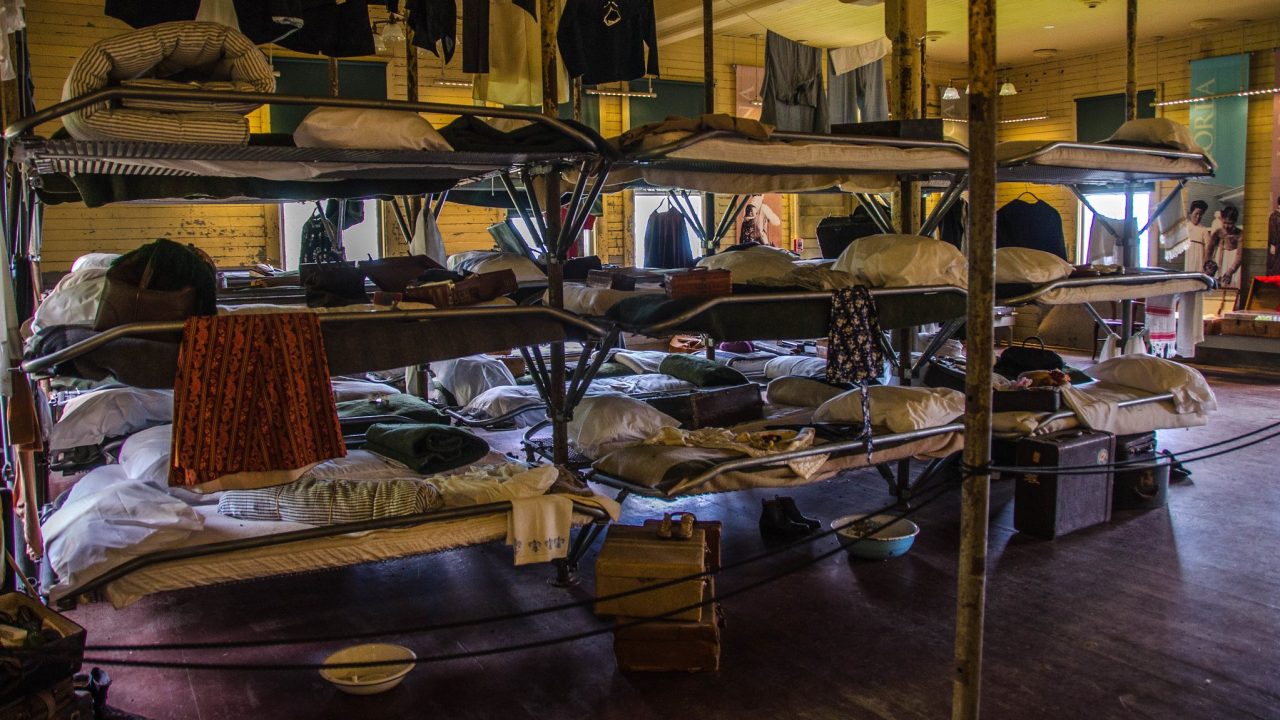 A row of bunk beds draped in clothing and other personal possessions, roped off inside a museum space.