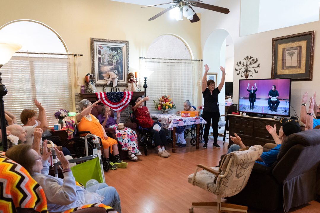 A group of seniors and senior care staff seated around a television showing a video of a drummer and instructor