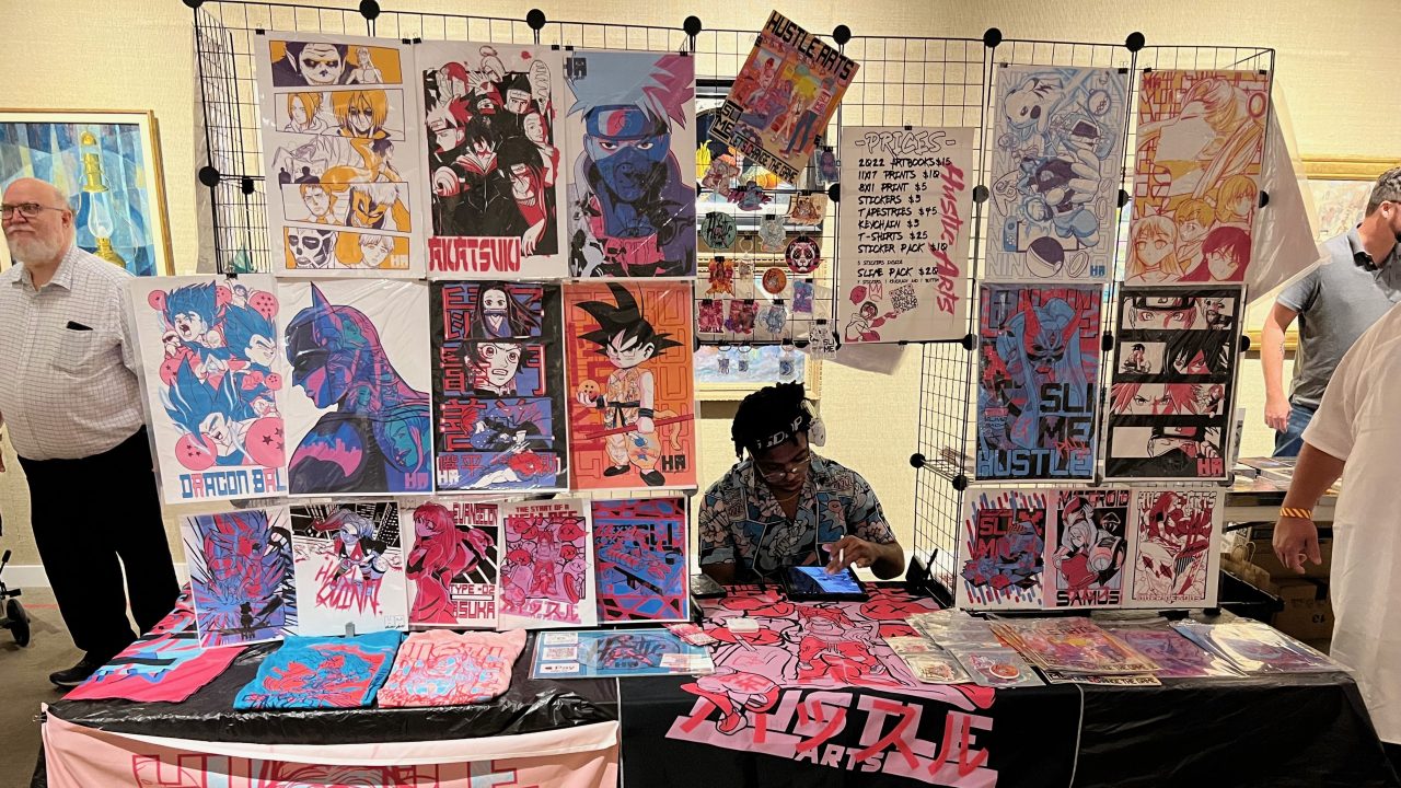 An artist sitting at a temporary booth set up in a museum gallery with anime-inspired drawings on display