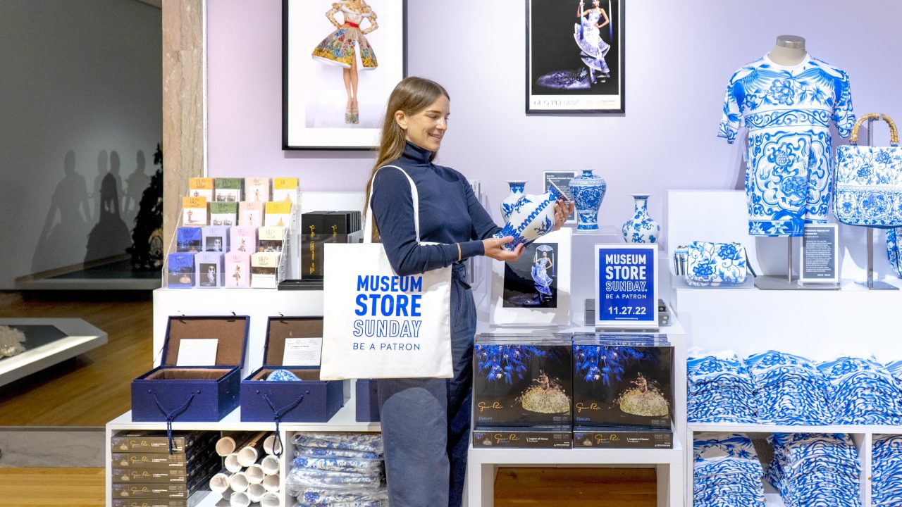 A person admiring a vase in a museum store while wearing a tote bag that says "Museum Store Sunday"