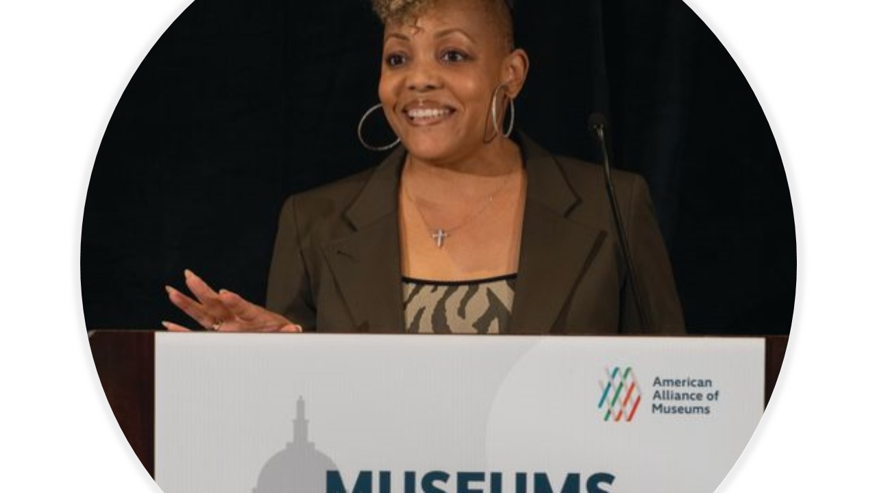 A woman stands smiling behind a podium with a sign on the front that says "Museums Advocacy Day" on the front.