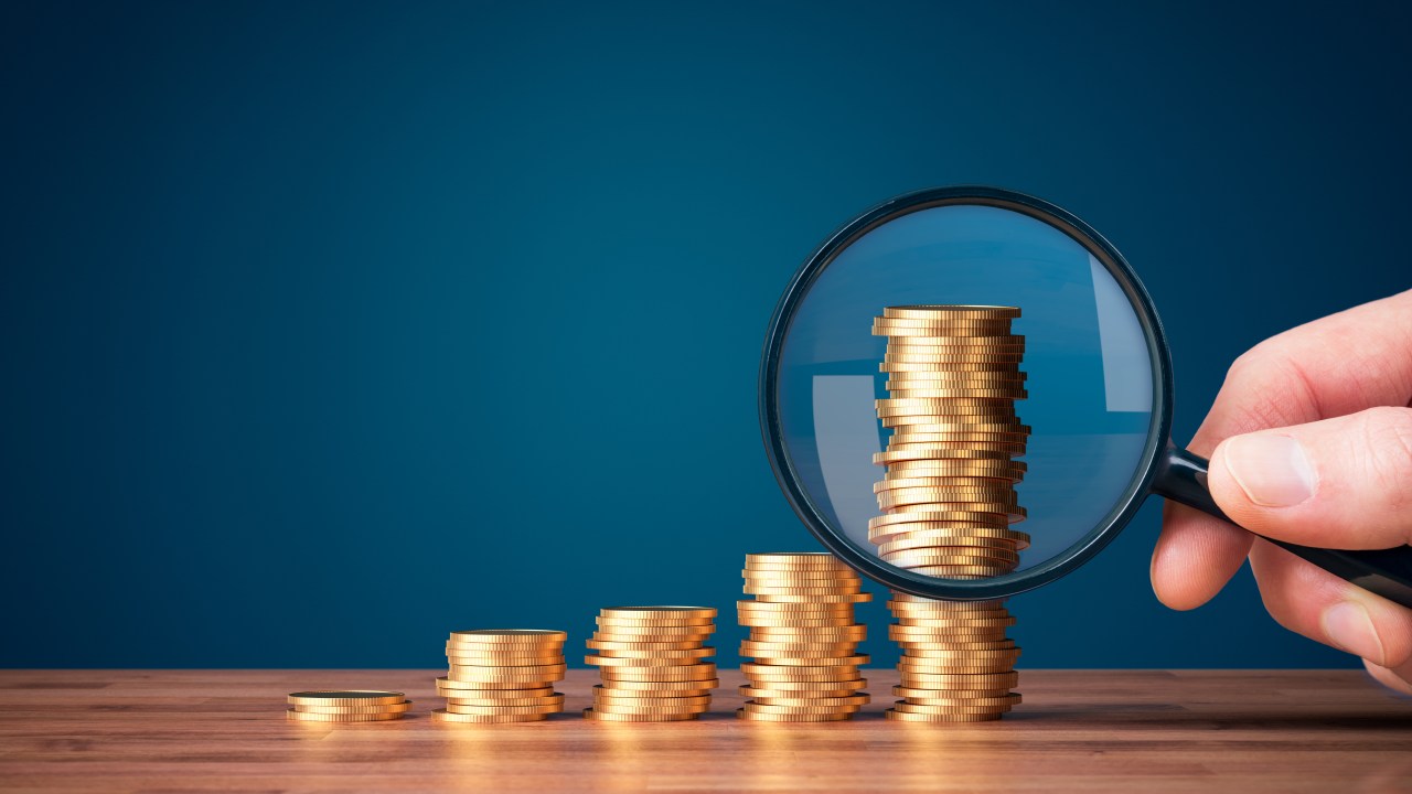 A hand holding a magnifying glass over stacks of gold coins.