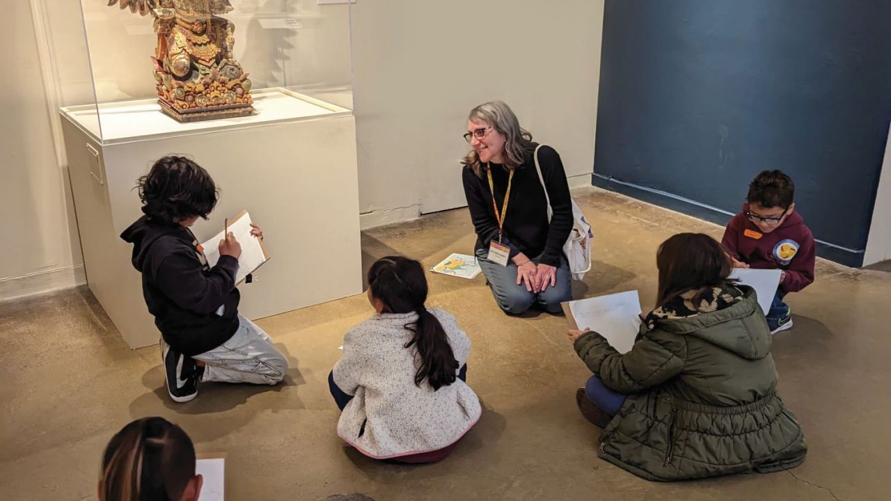 A gallery guide sits on the floor in an exhibition with a group of children scattered around listening.