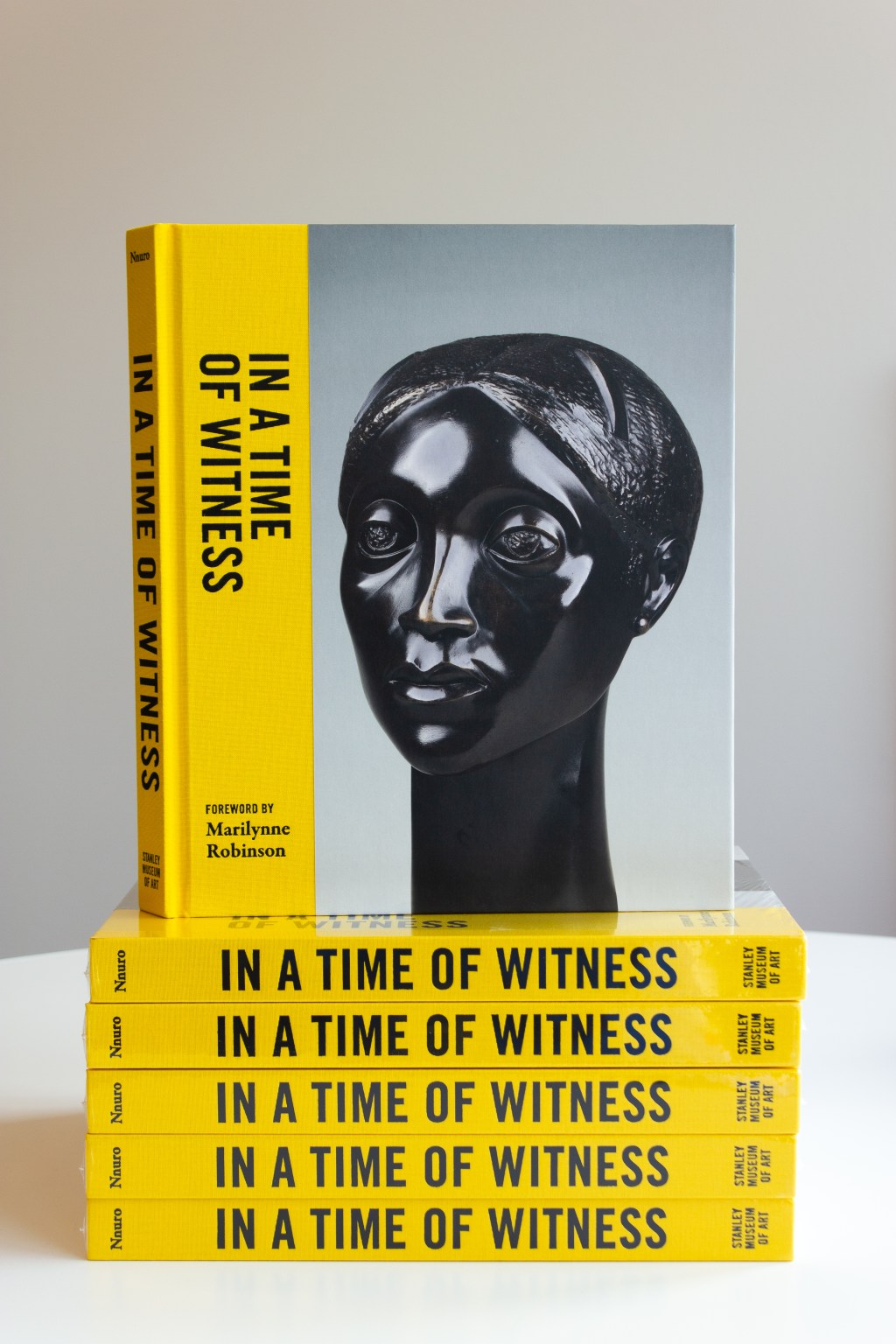 A stack of books with the title "In a Time of Witness" on the spine and cover, advertising a foreword by Marilynne Robinson and featuring a black sculpture of a human face