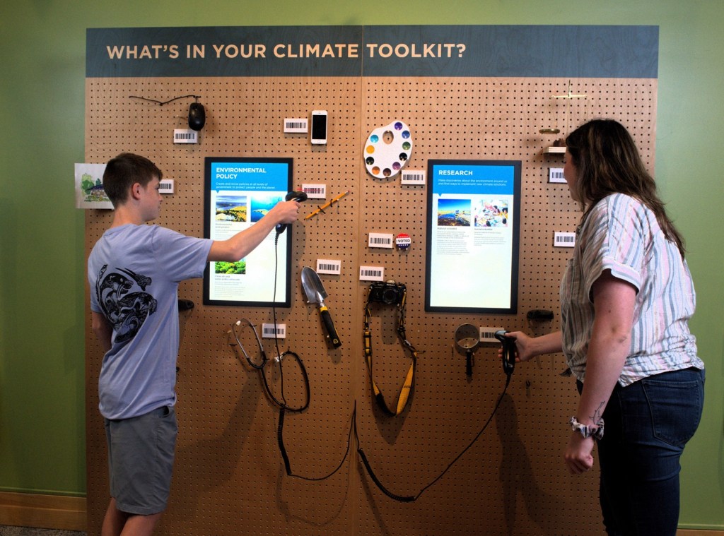 Two people stand at the Climate Toolkit interactive which includes two touchscreens and various artistic and other types of tools to manipulate the interactive with. 