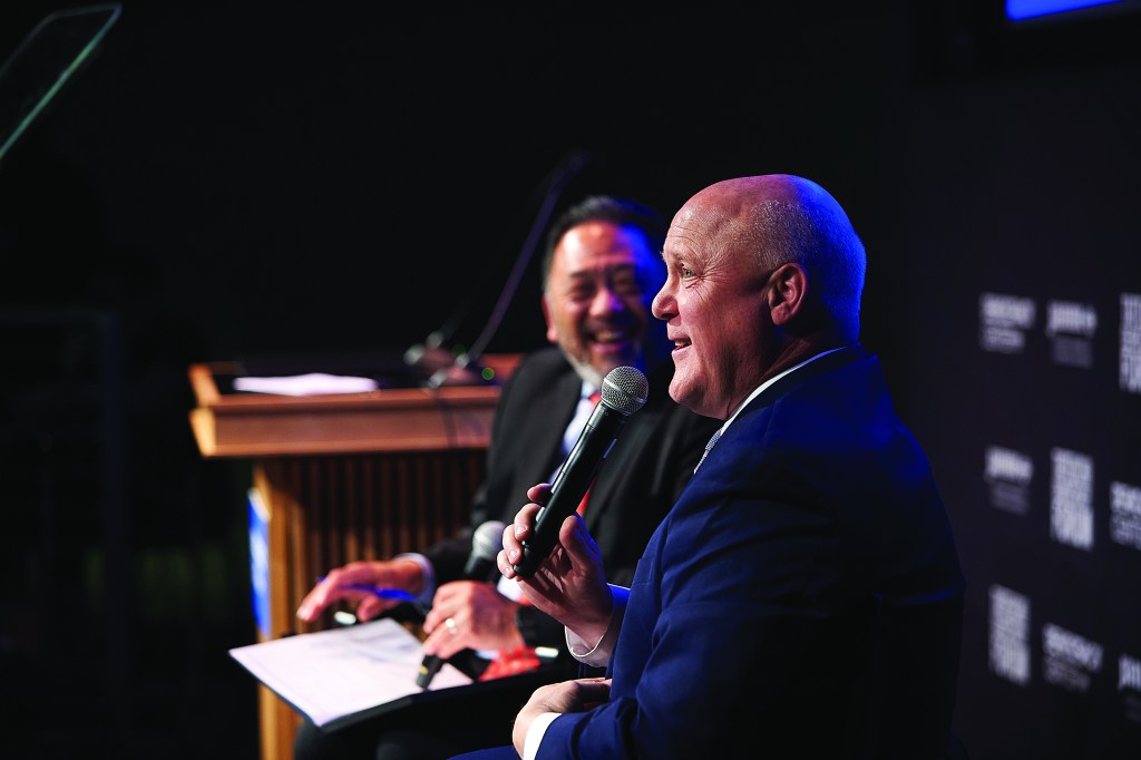 Two people sitting on a stage, talking to a crowd with a microphone