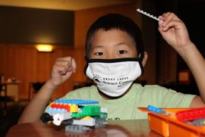 A young boy sits at a table with a Lego creation in front of him holding up a Lego piece in his hand. He is wearing a facemask over his mouth. 