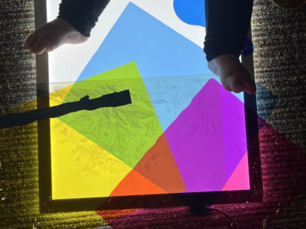 A baby sitting on a light table, with colorful transparent plastic sheets on the surface