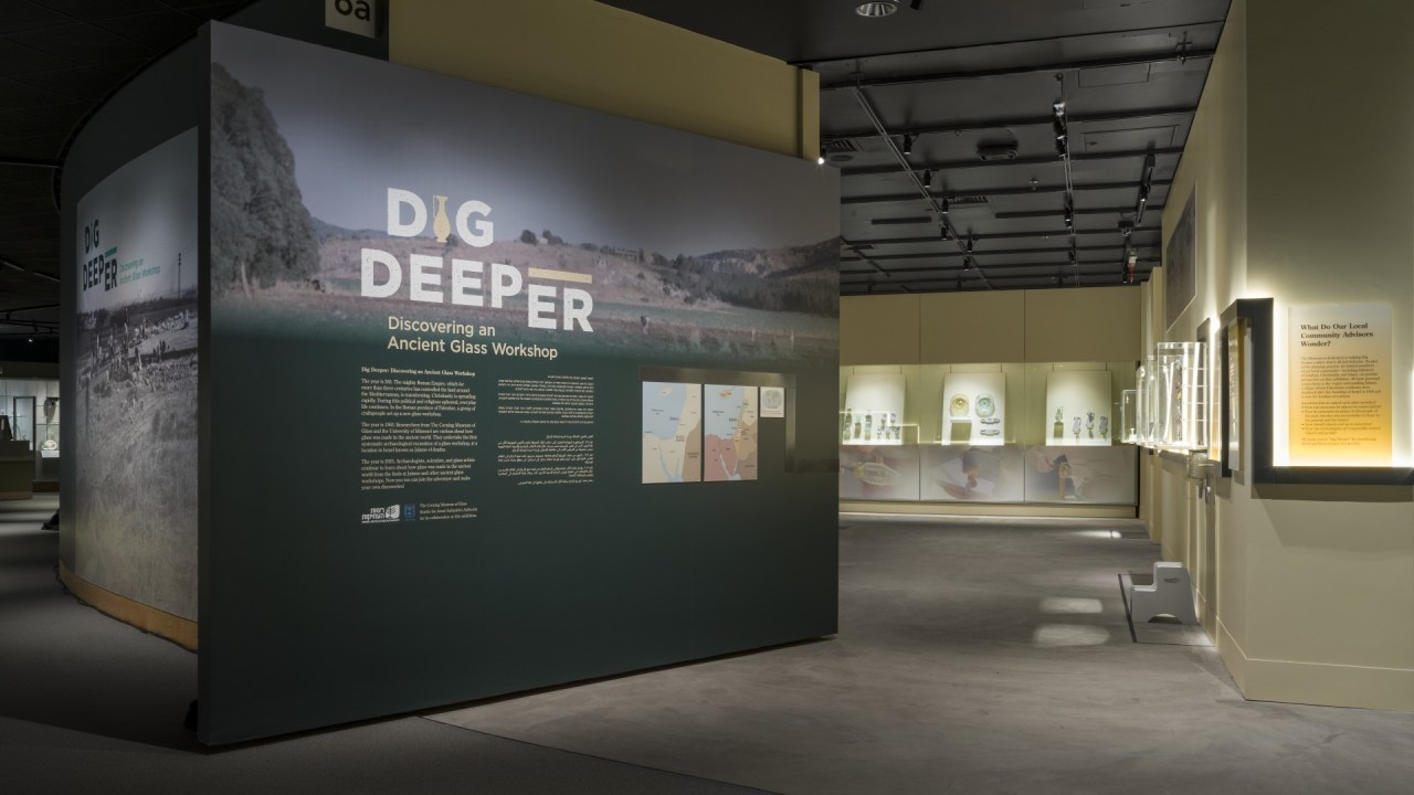 The entrance to the Dig Deeper exhibition with a slanted text wall in green and drawing visitors toward the right where cased objects are on display.