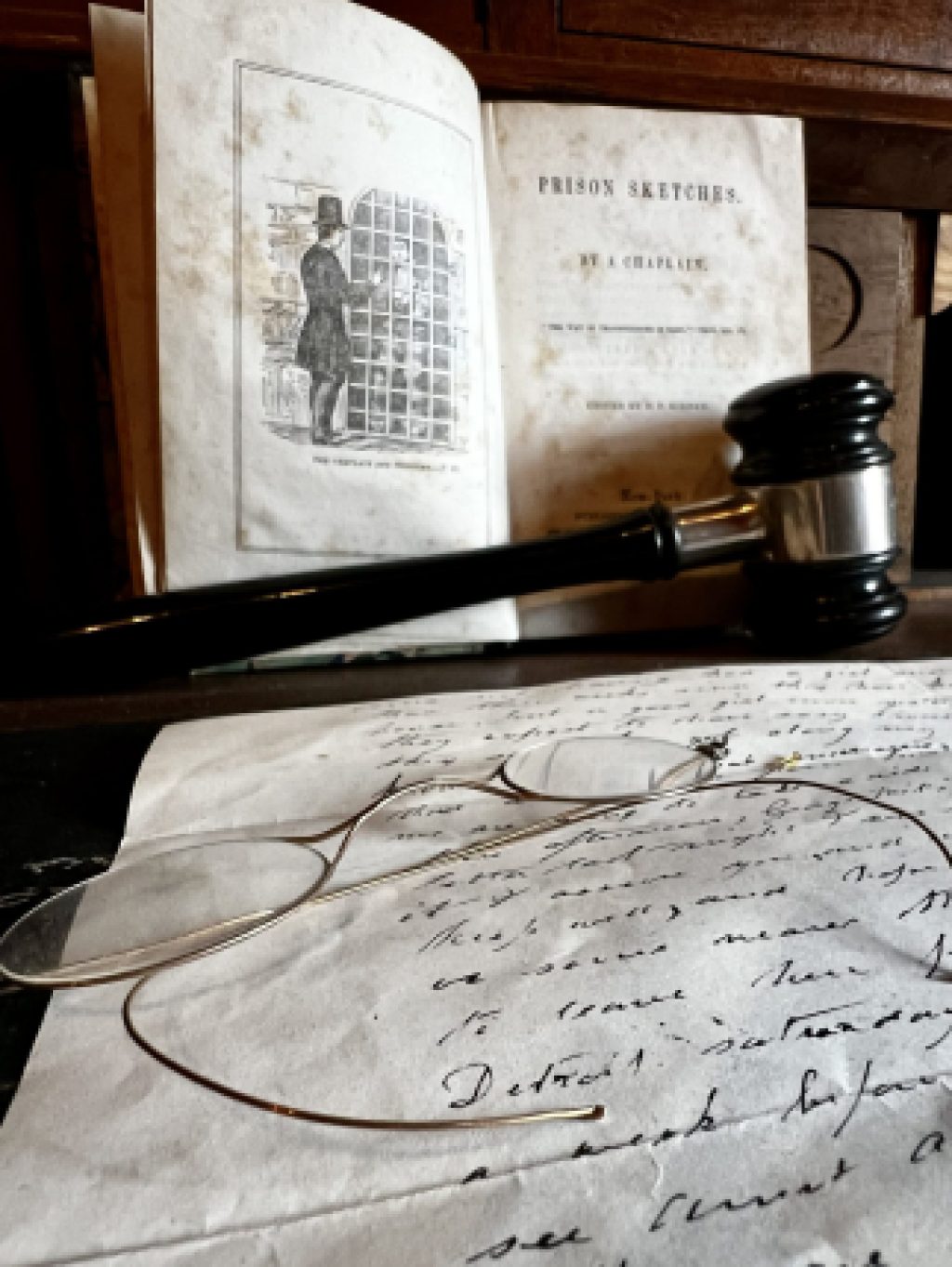 A closeup of a handwritten note with a pair of wire rimmed glasses sit on a table where an open book sits with a judge's gavel in front.