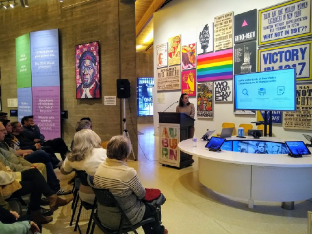 A small group of people sit in front of a podium where a person is speaking standing at the entrance to an exhibition.