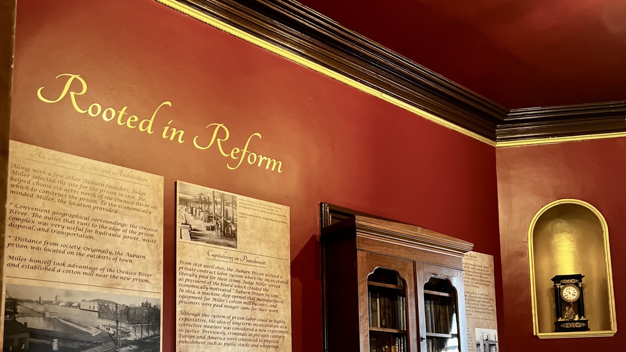 The interior of a historic house museum exhibition with a red wall with several text panels, a desk and chair in the corner near a fireplace on the next wall.