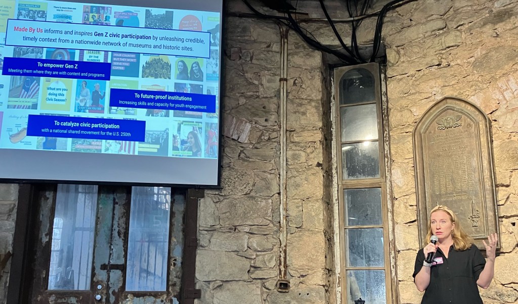 A speaker on a stage in a historical building in front of a slide reading "Made By Us informs and inspires Gen Z civic participation by unleashing credible, timely context from a nationwide network of museums and historic sites."