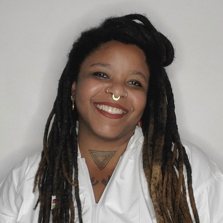 A black woman stands smiling in front of a white backdrop with long braided hair pulled partially into a topknot with a silver nosering wearing a white button down shirt.