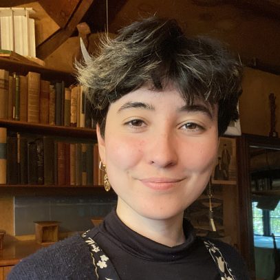 A white woman with short brown hair looks directly in the camera wearing a dark colored top and beaded necklace.