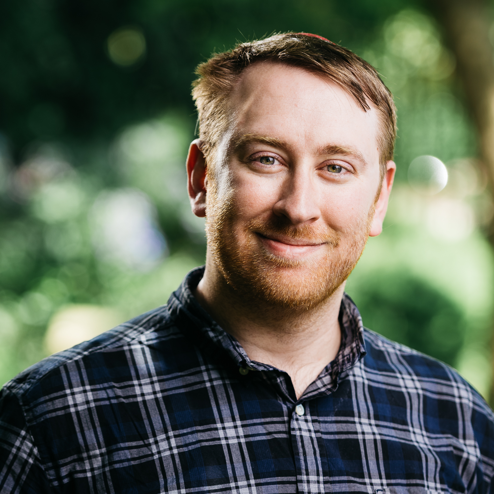 A bearded white man with light brown to reddish brown hair stands outside wearing a blue and white plaid shirt.