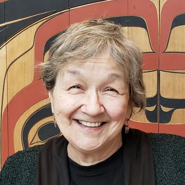 A white woman with short light colored hair stands in front of a wooden sculpture smiling.