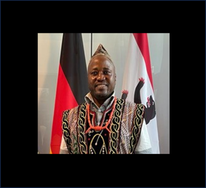 A black man stands in front of two different flags wearing traditional African garments.