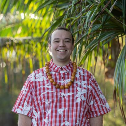 A native Hawaiian man stands outside with short dark brown hair wearing a red and white checkered and patterned short sleeve shirt and a Lai necklace.