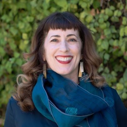 A white woman stands in front of greenery with long brown hair and short bangs wearing a dark teal colored top and scarf with long colorful earrings.