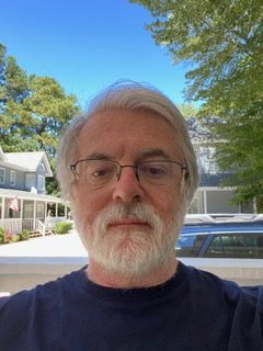 A bearded white man with silver hair and beard stands outside wearing a dark colored t-shirt.