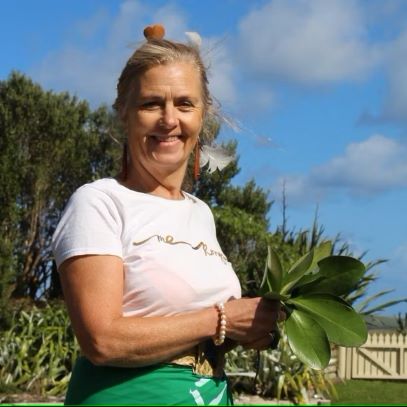 A white woman stands outdoors holding a plant with light colored hair pulled up on top of her head wearing a white t-shirt and green colored bottoms.