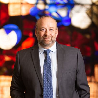 A bald white bearded man stands in front of a large stained glass window wearing a dark colored suit wiht a blue tie.