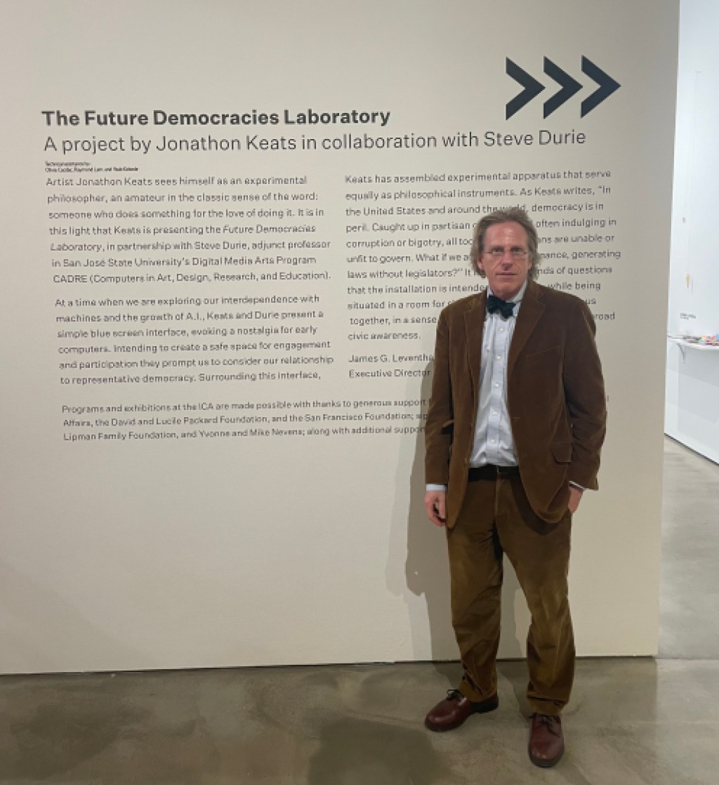 A person wearing a suit and bowtie in front of wall text panel reading "The Future Democracies Laboratory / A project by Jonathon Keats in collaboration with Steve Durie"