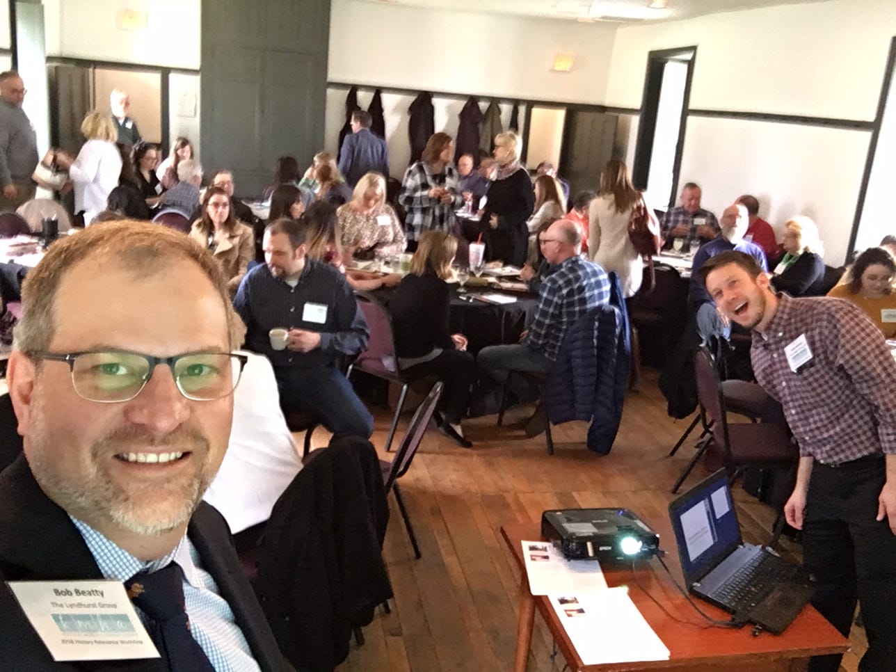 Bob Beatty takes a selfie image of the workshop room. He is in the bottom left corner of the frame wearing glasses with a large group of people seated around round tables behind him. 