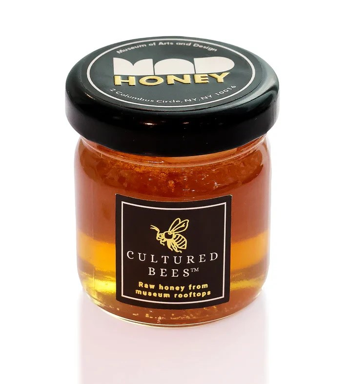 A jar of honey with a lid that reads "Museum of Arts and Design MAD Honey" and a label on the front that reads "Cultured Bees / Raw honey from museum rooftops"