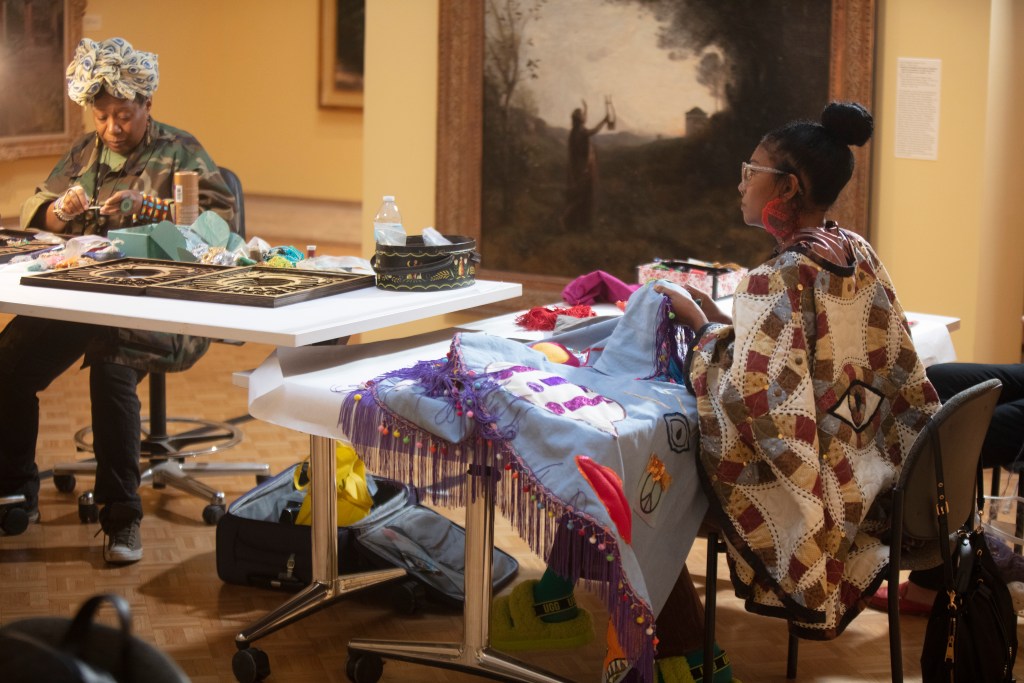 Two people working on textile projects on tables placed in a museum gallery