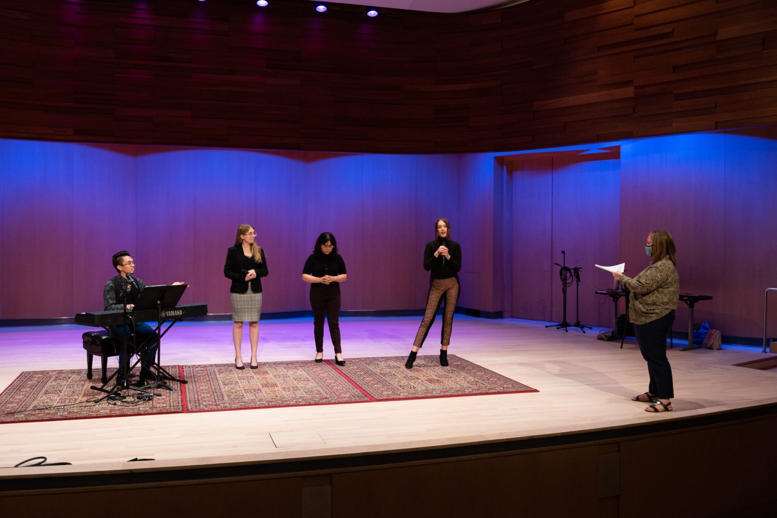 A group of performers standing on a stage with a director reading from notes to them