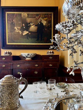 An ornately decorated dining room showing the edge of a tablecloth covered table with various glasses and dining ware a wooden sideboard in the background with a nicely framed painting of several gentlemen in the same room as shown. 