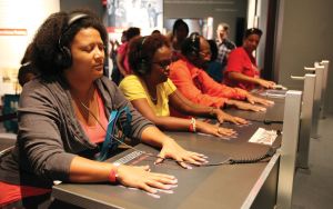 Image of a group of people sitting at a long tabel with their hands on hand prints in front of them each with a set of headphones listening. 